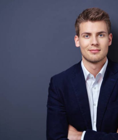 Confident handsome young businessman in a stylish jacket standing with folded arms smiling at the camera over a dark background with copy space
