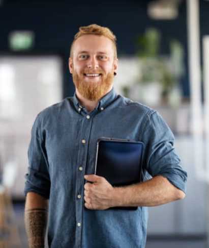 istockphoto-1265176370-612×612 Portrait of a businessman with beard standing in office holding digital tablet. Confident male business executive in office looking at camera.
