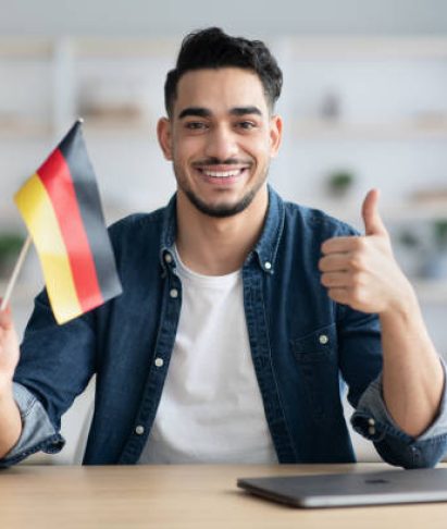 istockphoto-1322921058-612×612 Smiling arab guy with flag of Germany showing thumb up, sitting at desk with laptop, middle-eastern young man student learning German for job, education or emmigration to Germany, copy space